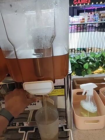 The video presents a close-up shot of a beverage dispenser in what appears to be a food service or restaurant setting. The dispenser is a dual-container unit, with each clear plastic container holding a brown-colored liquid. A hand is seen operating the lever of one of the dispensers, filling a clear plastic cup with the liquid. To the right of the dispenser, a tray holds a spray bottle containing a clear liquid. The background shows a blurred scene of people and retail displays, indicating the location is likely a public area. Text is visible on the dispenser, including the words "CAUTION" and a longer phrase, which reads "REFILL KHUSUS YANG PESAN OCHA", indicating language other than English.