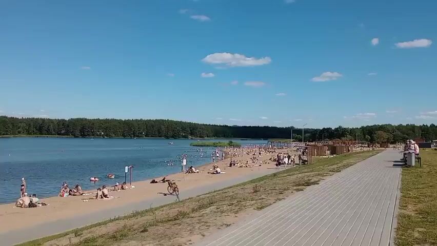 This media file captures a vibrant summer scene at a beach on a sunny day. The wide shot showcases a sandy beach teeming with people enjoying the warm weather. Individuals are scattered across the sand, some sunbathing, others engaged in conversations, and a few are in the water. The lake is calm, with a few people swimming or floating on inflatable toys. A paved walkway runs along the edge of the beach, with a few people strolling or sitting on benches.

The backdrop is a lush, green forest that lines the opposite shore of the lake. The sky is a brilliant blue, dotted with a few fluffy white clouds, suggesting fair weather. The time of day appears to be midday or early afternoon, given the bright sunlight and shadows.

The overall atmosphere is one of relaxation and recreation. People seem to be in good spirits, enjoying the day outdoors. The location is identified as Padlabenski Sel'ski Savet, Belarus. There is no visible text in the media file.