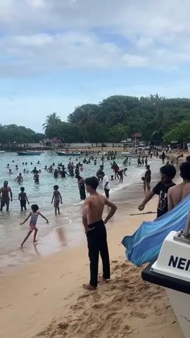 The scene captures a vibrant beach teeming with people enjoying the water. The beach features light brown sand, and the water extends into a bay surrounded by lush green trees and a protective wall.  Numerous individuals are submerged in the water, wading, swimming, and generally enjoying the sea. Children can be seen playing near the shore, with their guardians close by.

On the sandy beach, people are standing, walking along the shoreline, or simply observing the activities. A few people are conversing, adding to the lively atmosphere of the beach. A blue and white striped cloth is draped on a boat, visible at the edge of the frame.