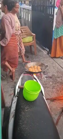 A woman is frying food in a pan over an open fire. She is using a spatula to turn the food in the pan. Another woman is standing to her left, and a third woman is standing to her right. There is a green bucket on the seat of a motorcycle in the foreground. The scene appears to be outdoors, possibly on a street or in a courtyard.