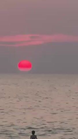 A young person stands in shallow ocean water facing the sunset. The sunset has a large red sun over the horizon, with light pink and gray sky above it. Gentle waves lap around the person. The person's back is to the camera.