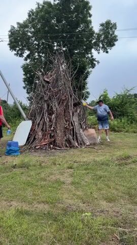 Two men stand in a grassy area with a large pile of wood in the center, likely for a bonfire. A tall tree is positioned behind the woodpile. The man on the right, wearing a blue t-shirt and shorts, is holding a yellow bottle and appears to be pouring liquid onto the wood. The man on the left, wearing a red t-shirt and shorts, is holding a blue bucket and a white object, possibly a sheet of metal. The sky is overcast.