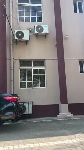The video captures a static shot of the exterior of a building in Ijanikin, Nigeria, during the daytime. The building features a light peach or beige-colored facade with a dark brown lower section. A prominent dark brown vertical column runs along the right side of the building, extending from a white circular base on the ground.

Two air conditioning outdoor units are mounted on the upper portion of the wall. The unit on the right has the brand name "Hisense" visible on it. Another AC unit is mounted slightly below and to the left of the Hisense unit. In the central part of the frame, there is a large, white-framed window divided into multiple panes, reflecting trees and foliage. A small white light fixture is mounted on the wall above the brown column.

At the bottom left, the rear section of a black car is partially visible. Below the car, near ground level, another smaller AC outdoor unit is enclosed within a metal cage. The ground in the foreground is paved, appearing to be interlocking stones or asphalt. No people are visible, and no specific activity is taking place; the video presents a still view of the building's exterior.