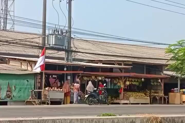 A street scene depicts a fruit stand in front of a long, low building with a corrugated metal roof. The building appears to be a covered market or shop with an open front. Several people are visible at the fruit stand, some appearing to be customers and others vendors. Various fruits, including what look like durians and coconuts, are displayed. A red and white Indonesian flag is mounted on a pole to the left of the stand. Multiple motorcycles are present, with some individuals riding them and others parked. A white van is driving on the road in front of the stand. The environment is an outdoor setting with power lines and utility poles overhead. The sky is clear and bright, suggesting daytime.