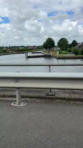 A first-person perspective video pans across a canal. A metal guardrail runs across the bottom of the frame, separating the viewer from the canal. The canal is wide with calm water. On the far bank, several houses and buildings are visible, nestled amongst trees. A lock system with gates is present in the middle of the canal. A boat can be seen entering the lock system. The sky is partly cloudy with white clouds against a blue sky. The overall scene depicts a tranquil waterscape with hints of human activity.