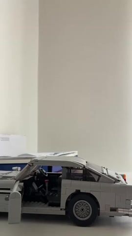 A white Lego car is shown on a table. The driver's side door is open, revealing the interior of the car. A person's hand reaches into the frame and closes the door. The person's hand then reaches to the rear of the car and opens the trunk. The trunk lid is raised. The video is shot from a side angle, with the car in the foreground and a white wall in the background.
