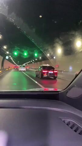 The video shows a view from inside a car traveling through a tunnel. The car's dashboard is visible in the foreground. The tunnel is illuminated by overhead lights, some of which appear to be green. Several other vehicles are visible on the road ahead, including a dark-colored sedan directly in front. The sedan's brake lights are illuminated. The road surface appears wet.