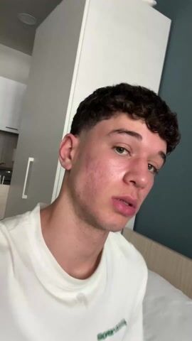 A young man with curly brown hair is the focus of the video. He has a fair complexion and green eyes. He's wearing a white t-shirt with green text. The camera appears to be handheld, likely a selfie. The background reveals a simple apartment interior. There's a wardrobe, a bed with a light wooden headboard, and a glimpse of a kitchen area with white cabinets. The man runs his hand through his hair at one point in the video.