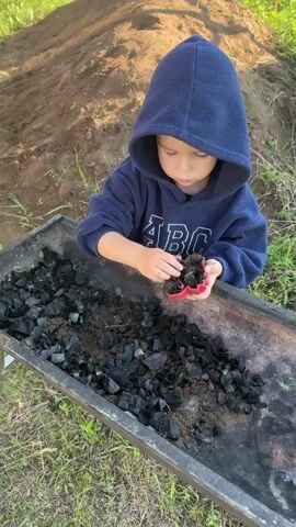A young child wearing a blue hooded sweatshirt with "ABC" written on it is interacting with charcoal and dirt in a dark-colored metal container, possibly a grill or a bin. The child holds a red object, which appears to be a heart-shaped cookie cutter, filled with charcoal and dirt. The child is looking down at the object in their hands. The background shows a dirt pile and green grass, suggesting an outdoor setting. Another child can be seen in the background to the left, holding an orange shovel. The lighting suggests it is daytime.