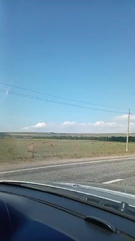 This video shows a view from inside a vehicle driving on a paved road. The road is in the foreground, with a white line marking the lane. To the right of the road, there is a utility pole with wires extending across the frame. Beyond the road, there is a large, open field with scattered hay bales and a few scattered trees. In the distance, there are rolling hills under a clear blue sky with a few white clouds. The scene appears to be rural and possibly agricultural. The video is shaky, indicating the vehicle is in motion.