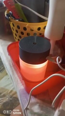 The video shows a humidifier with a black top and a translucent, illuminated cylinder base. The humidifier is placed inside a red plastic tray on a patterned surface. The light from the humidifier cycles through different colors, starting with orange, then turning yellow, and finally green. A white power cord is connected to the humidifier. In the background, a yellow plastic basket containing hair accessories, such as combs, is visible. There is also a white object, possibly another bottle or container, next to the humidifier. The scene appears to be indoors, possibly on a table.