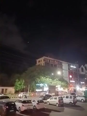 The video captures a nighttime scene in what appears to be a city area. The sky is dark, with some clouds visible.  A few lights are visible in the distance, possibly from an aircraft. Below the sky, several buildings can be seen. One is a multi-story structure with a light-colored facade and multiple windows. There are trees in front of the building, partially obscuring the lower levels.  The street is lined with parked cars of various makes and colors, including several white hatchbacks. In the middle of the street, people can be seen near the parked cars. The overall scene appears to be a busy urban environment at night.