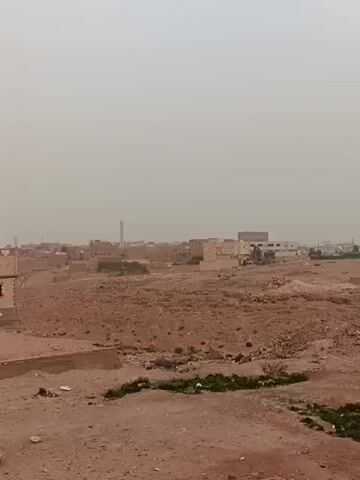 The video shows a landscape consisting of a dusty, reddish-brown terrain with scattered rocks and sparse vegetation. In the distance, a small town with light-colored buildings is visible. The sky is overcast, contributing to a hazy, muted light. The camera is static, providing a wide, establishing shot of the scene.
