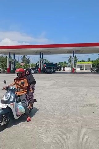 The video shows a person riding a scooter out of a gas station in what appears to be a sunny, tropical location. The rider is wearing a red helmet and an orange shirt and has a passenger wearing a black hijab on the back of the scooter. In the background, there is a Pertamina gas station, other vehicles, and houses and trees. The video follows the scooter as it drives out of the station.