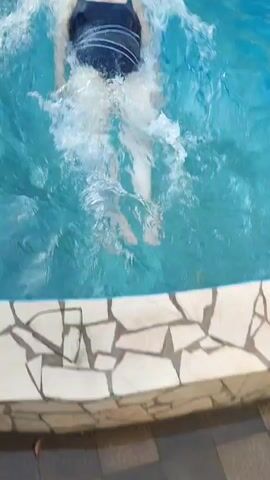 A person wearing a dark swimsuit floats on their back in a swimming pool. The person's arms are extended to their sides, and their legs are submerged in the water. The water is disturbed, creating ripples and splashes around the person. The pool is surrounded by greenery and a stone edge. The video is shot from a high angle looking down at the person.