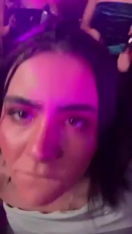 The video shows a person with dark hair and fair skin, wearing a light green dress, standing in a crowded place, possibly a club, with people around. The lighting is predominantly purple and blue. The camera is close to the person's face, and they are looking directly at the camera. The person seems to be moving or dancing to the music.