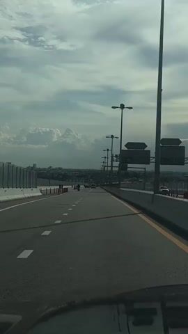 The video shows a POV shot from a vehicle driving on a highway. The highway is elevated, with a barrier on the right side and a pedestrian walkway on the left, separated by a fence. The road is paved and has lane markings. Several vehicles, including cars and motorcycles, are visible ahead, moving in the same direction. Streetlights are spaced along the right side of the highway. In the distance, a cityscape can be seen under a cloudy sky with large cumulus clouds. The overall impression is that of a typical highway scene on a day with somewhat overcast weather.