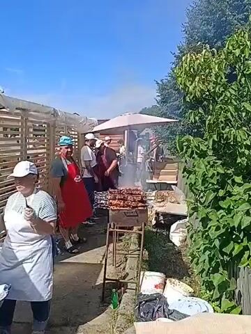The video shows an outdoor gathering with several people, likely a barbecue or festival. In the foreground, a woman wearing a white apron and a white cap is preparing food at a green table with cooking equipment, including a portable stove and pans. She is holding what appears to be a piece of dough or a small patty. Behind her, to the left, is a wooden structure with slatted walls. In the center, there is a grill with meat cooking over flames, generating smoke. Several individuals, mostly men, are standing around the grill and a nearby table. Some are wearing aprons. Behind the grill is a large umbrella providing shade. The background features trees and a clear blue sky. Trash bags and various containers are visible on the ground. The overall impression is a casual outdoor event focused on food preparation and consumption.