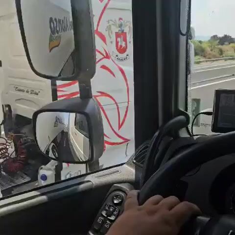 The video is filmed from the perspective of someone driving a truck. The driver's hands are visible on the steering wheel, which has several buttons. A GPS device is mounted on the dashboard. Outside the truck, other vehicles, including another truck, are visible on a highway. The weather appears to be clear. The video includes a series of short clips, each showing a slightly different view inside the truck's cab or of the road ahead.