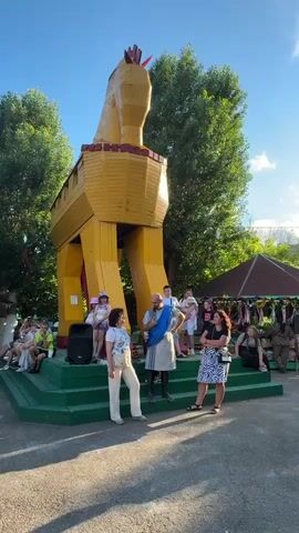 The video captures an outdoor gathering centered around a large, yellow, wooden replica of the Trojan Horse. A man dressed in a toga-like garment and a blue sash stands on a green platform in front of the horse, speaking into a microphone. He appears to be addressing a small group of people, some of whom are seated on benches to the left and right of the platform. The people in the audience are a mix of adults and children, casually dressed. To the right of the platform, a stall with various items, possibly souvenirs or crafts, is visible, suggesting a market or fair-like atmosphere. Trees and a clear blue sky form the backdrop. The overall scene suggests a historical reenactment or a themed event.