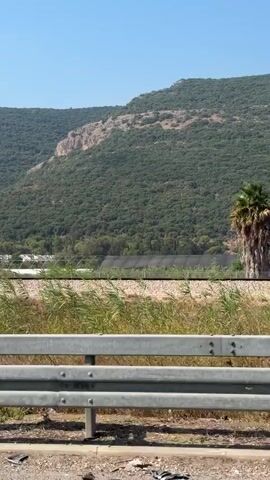The video shows a view from a moving vehicle, looking out at a rural landscape. In the foreground, there is a metal guardrail, and beyond that, a field of tall, green grass. Further in the distance, there are several white-domed structures, possibly greenhouses, situated in a valley. The background is dominated by rolling green hills covered in trees, with a clear blue sky above. The sun is shining, casting shadows and illuminating the scene. There are no people visible in the video.