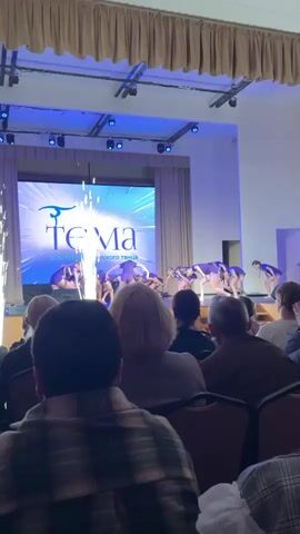 The video shows a stage performance in front of an audience. A group of girls, wearing matching blue and purple outfits, are on stage standing up after posing at the beginning of their routine. Fireworks are shooting up from the floor on either side of the stage. The screen in the background is blue and reads “Tema” in Russian. The audience sits in the foreground with their backs to the camera. The person in the very front is wearing a plaid shirt. One person holds up their phone to take a photo. A stagehand wearing a gray outfit runs onto stage carrying what looks like a clipboard. The stage has a dark brown curtain on top.