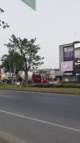 A red cement mixer truck is parked on the side of a road. It is facing right, with the drum angled slightly upward. A young tree with a supporting stake is located to the left of the truck. In the background, there is a row of buildings with various signs, including one that says "Kopi Kenangan" with a heart symbol. The sky is overcast.