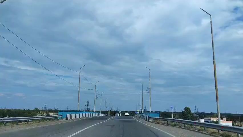 A first-person perspective video captures a drive across a bridge on a cloudy day. The road has two lanes in each direction, separated by a white line. Blue and white striped barriers line the sides of the bridge, with metal railings on top. Several streetlights and power lines are visible along the route. Two cars are ahead of the camera, one black and one white. The landscape on either side of the bridge consists of flat terrain with some vegetation and distant structures. The sky is filled with dramatic, fast-moving clouds.