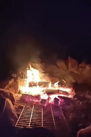 This is a short video clip showing a campfire at night in Būtkiemis II, Lithuania. The fire is burning brightly with flames reaching upwards, casting a warm glow on the surrounding logs and rocks. A metal grate sits in front of the fire, suggesting the possibility of cooking. The scene is dark, indicating it's nighttime, and there's a subtle hint of smoke rising from the fire. No people are visible, but the presence of the grate and the implied gathering around a campfire suggest a recreational or social setting, likely outdoors. The overall mood is serene and cozy, with the crackling fire providing a natural focal point. There is no visible text in the video.