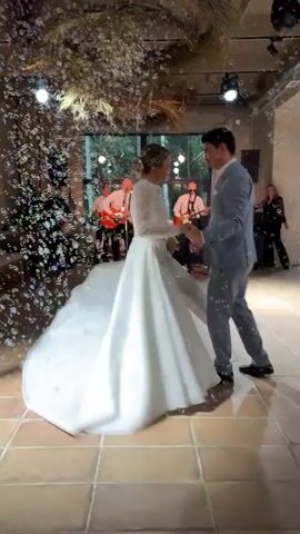 A wedding reception is underway. In the foreground, a bride in a white wedding dress and a groom in a light blue suit are dancing. They are surrounded by a stream of bubbles. In the background, a live band with two guitarists and a drummer is performing on a stage. A woman is visible standing near the stage. The room has tiled floors and exposed beams on the ceiling, with decorative dried foliage hanging down. Lights are visible on the ceiling.