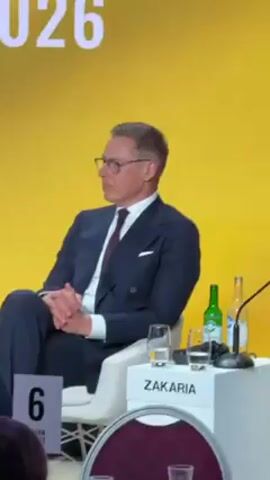 The video shows a man in a dark blue double-breasted suit, a white shirt, and a dark red tie, sitting in a white chair on a stage with a yellow backdrop. He is wearing glasses and has his hands clasped in front of him. A name card with the name "ZAKARIA" is visible on a small white table next to him, along with a glass of water, a bottle of green liquid, a bottle of clear liquid, and a microphone. A number "6" is displayed on a small sign near the front of the stage.