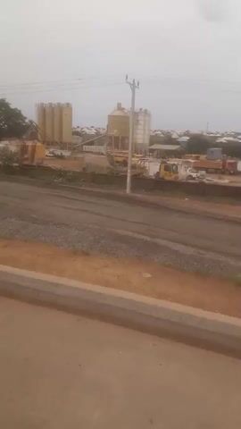 The video shows a construction or industrial site under a cloudy sky. There are several large storage tanks, piles of gravel, and multiple dump trucks. A few trees are also visible. The camera moves, revealing more of the industrial area, with what appears to be a residential area visible in the background.