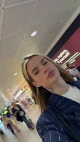 A woman is walking through an airport terminal. She is wearing a navy blue pinstripe blazer, a white shirt, and a silver necklace. She has blonde hair with a light blonde streak at the front. She is pulling a small, dark suitcase with a blue neck pillow attached to the handle. The airport terminal has a white ceiling with recessed lights and light grey tiled floors. There are glass walls on one side of the corridor and a travelator in the middle. People are walking in both directions. There is a sign indicating the way to gates G35-G48 and G51-G54.