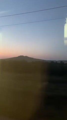 The video shows a view from a moving vehicle, likely a train, looking out a window. The time of day appears to be either sunrise or sunset, with a pale blue and pink sky. In the distance, a large, dark, undulating hill or mountain range is visible. The foreground is blurred, suggesting movement, and consists of dark shapes that could be trees or foliage, and a bright, hazy area on the left side of the frame, possibly from the sun or reflections. A reflective surface, possibly the window itself or another part of the vehicle's interior, is visible on the right side of the frame, showing a rectangular white surface. A thin, dark horizontal line, likely a power line or wire, runs across the top of the frame. The overall impression is a scenic, fleeting glimpse of a landscape in motion.