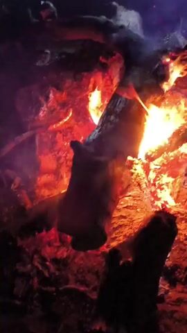 The video shows a close-up view of a campfire at night. Large logs are burning, with flames and embers visible. Smoke rises from the fire. The ground around the fire appears to be dirt or ash. The lighting is dim, with the fire providing the primary light source, casting a warm, orange glow on the surroundings. There are no people visible in the frame. The camera angle is slightly from above, looking down into the fire.