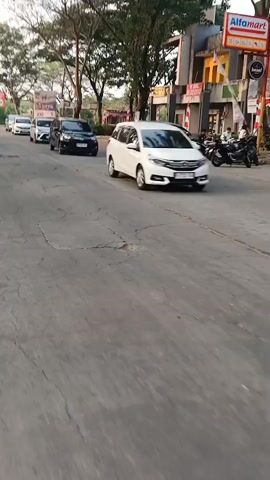 The video shows a street scene with several cars driving and parked, including a white Honda Mobilio, a black car, and a silver car. There are also trees and buildings lining the street. A person on a motorcycle is visible in the foreground, and there are a few pedestrians in the background. The road surface is asphalt and appears to be in fair condition with some cracks. A sign for Alfamart is visible on a building. The sky is overcast, suggesting it might be late afternoon or early evening. The general impression is a suburban or commercial street in an urban area.