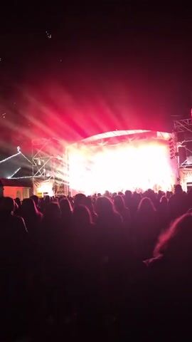 A crowd of people is standing in front of a concert stage at night. Fireworks are going off above the crowd and to the left of the stage. The stage is brightly lit with white and red lights, creating beams that shine through the crowd. In the background, a church with a cross on top is visible, decorated with lights. The camera remains mostly still.