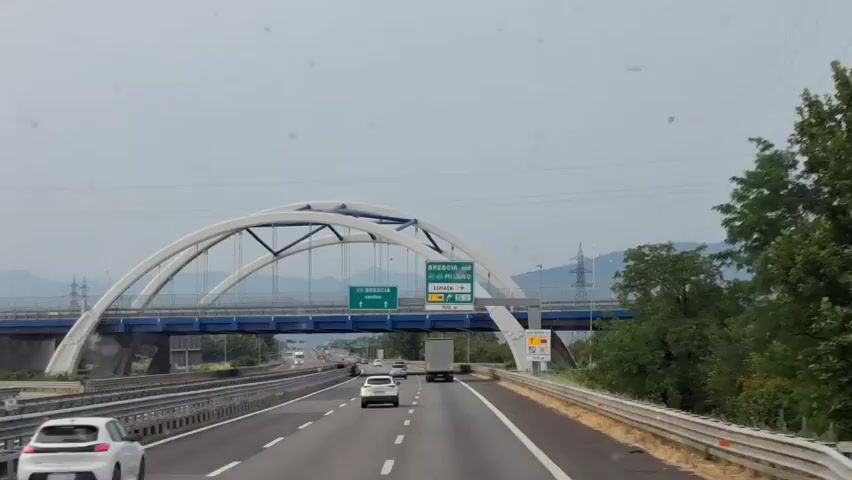 A highway scene in Italy. Cars are driving on the highway. There is a bridge in the distance with signs. The weather is overcast. The video also shows a view in the side mirror of a car driving on the highway. The side mirror reflects the highway and fields of crops. The sun is visible in the sky.
