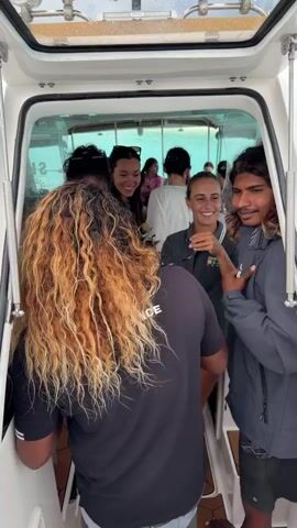 Inside a white boat cabin, a group of people gather near the entrance. Several individuals can be seen, including one with long, wavy blonde and brown hair facing away from the camera. Others are visible within the cabin, some wearing sunglasses and casual clothing. One person is holding up a pair of glasses, seemingly in jest. The interior of the cabin appears bright, with light reflecting off the windows. Some individuals are wearing clothing with the Maldives flag on it. The overall atmosphere seems relaxed and jovial.