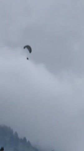 A paraglider descends through a cloudy sky. The paraglider is a dark-colored canopy with a person suspended beneath it. Below the paraglider, the top of a tree-covered hillside is visible. The sky is overcast with thick, gray clouds. The footage is shaky and appears to be handheld.