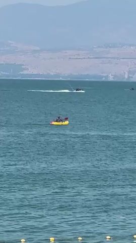 A body of water, likely a lake, with a gentle ripple pattern. In the mid-ground, a yellow kayak with two individuals is moving from left to right across the frame. One person in the kayak is holding a paddle. Further in the distance, a jet ski with two people is creating a white wake as it moves from left to right. In the far background, a mountainous shoreline is visible. The sky is overcast.