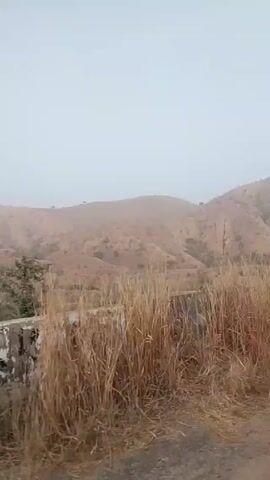 The video appears to be shot from a moving vehicle driving on an asphalt road winding through a hilly, arid landscape. The sky is overcast and visibility is low, giving the scene a hazy appearance. The hills are covered in dry vegetation, with sparse trees dotted across the landscape. A concrete barrier with graffiti lines the edge of the road. The road itself is cracked and shows signs of wear. The camera angle is from the driver's perspective.