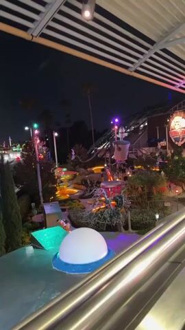 The video, taken at night, begins with a high-angle view of a brightly lit miniature golf course featuring alien-themed structures, next to a highway with moving cars. The camera shifts to the right, revealing a street with palm trees and streetlights. The video ends with a view of the road, lined with manicured greenery and illuminated by streetlights, and with the faint glow of other buildings in the distance.