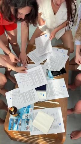 Several people stand around a rectangular coffee table covered with documents, a smartphone, a passport, and travel brochures. The documents on the table have blue dots on them. Some of the people are holding documents in their hands and reading them. A woman in a red outfit is pointing at an object on the table. The coffee table is situated on a tile floor. The scene is shot from an overhead perspective.