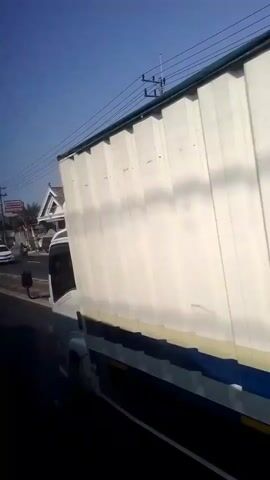 A slightly shaky, wide-angle video appears to be filmed from the window of a vehicle traveling on a road. The perspective is low, showing the side of a white truck in the foreground. Beyond the truck, a street is visible with several buildings lining one side. One building, painted yellow and white, has a golden gate and appears to be a shop or business. Further down the street, a convenience store with red and yellow signage is visible. Other vehicles, including a white car and a red SUV, are seen on the road. The sky is clear and blue. The video captures a brief glimpse of a street scene in what appears to be a commercial or residential area with some traffic.