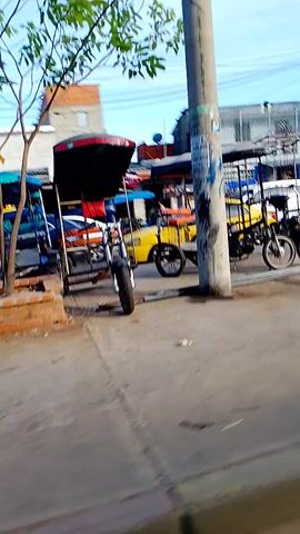 The video shows a street scene with a yellow tricycle taxi parked alongside a white van. The buildings in the background are a mix of brick and painted structures, with visible power lines overhead. A silver car is also parked along the street. The camera appears to be in motion.
