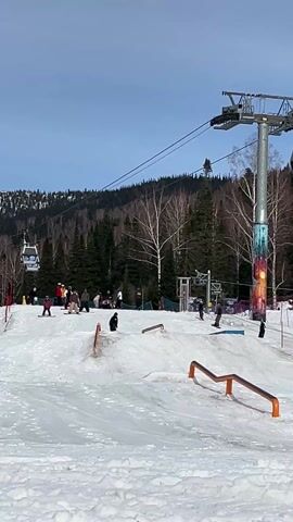The video shows a snowy ski area, possibly a terrain park, with several individuals engaged in skiing and snowboarding activities. In the background is a ski lift operating with visible cabins moving along the cables. The area is punctuated with orange metal rails designed for skiers and snowboarders to perform tricks. In the foreground, two children wearing yellow vests and helmets are standing on skis. In the background, a snowboarder is attempting to maneuver on one of the rails. The weather is sunny and the sky is mostly clear. Trees and a distant mountain are also visible.