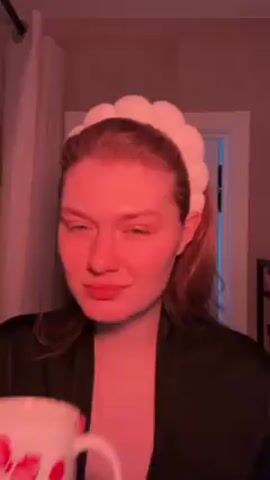 A fair-skinned woman with reddish hair, wearing a black robe and a white, puffy headband, drinks from a Hello Kitty mug. She's standing in what appears to be a bathroom. The lighting is predominantly red, but a doorway in the background is illuminated in blue. The woman takes a sip, then tilts her head back and looks up.