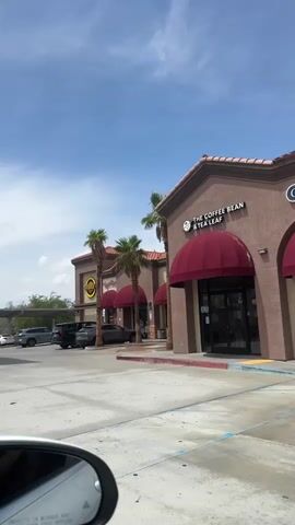 The video pans across the exterior of a small strip mall on a sunny day. Palm trees line the front of the buildings. The prominent storefronts visible are The Coffee Bean & Tea Leaf, and another business with a circular yellow sign that appears to be a restaurant. Several cars are parked in front of the businesses. The view is from a car, with a portion of the driver's side mirror visible in the foreground, showing the reflection of the sky and a building. The sky is blue with scattered white clouds. The ground in front of the strip mall is paved concrete.