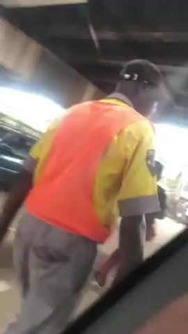 The video was recorded from inside a vehicle moving under a bridge. The environment is a busy street with pedestrians, vehicles, and commercial structures. The first individual seen is wearing a yellow shirt, an orange vest, and gray pants. They walk towards the vehicle recording the video. Then, a person in a dark shirt and red pants is seen walking away from the camera's perspective. In the background, there is a yellow taxi, other vehicles, and people milling around. The overall scene suggests a bustling urban environment.